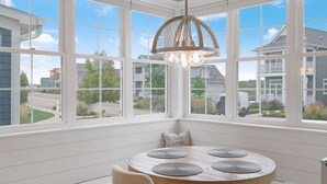 A room with a large bay window, a round table with placemats, a hanging light fixture, and a pillow with text. - Sit, Stay, Relax | Welcome to Serendipity (Ottawa)
