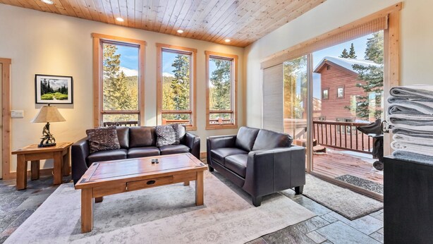 A cozy living room with a wooden ceiling, large windows, a fireplace, and a view of a cabin. - Mountain Retreat with Deck and Hot Tub (Blue River)