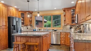 A spacious kitchen with wooden cabinets, a large island, and a view of a house through a window. - Mountain Retreat with Deck and Hot Tub (Blue River)