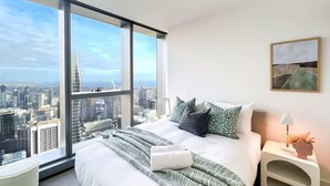 Une chambre moderne avec un grand lit, une table de chevet et une vue sur le paysage urbain grâce à de grandes fenêtres du sol au plafond.