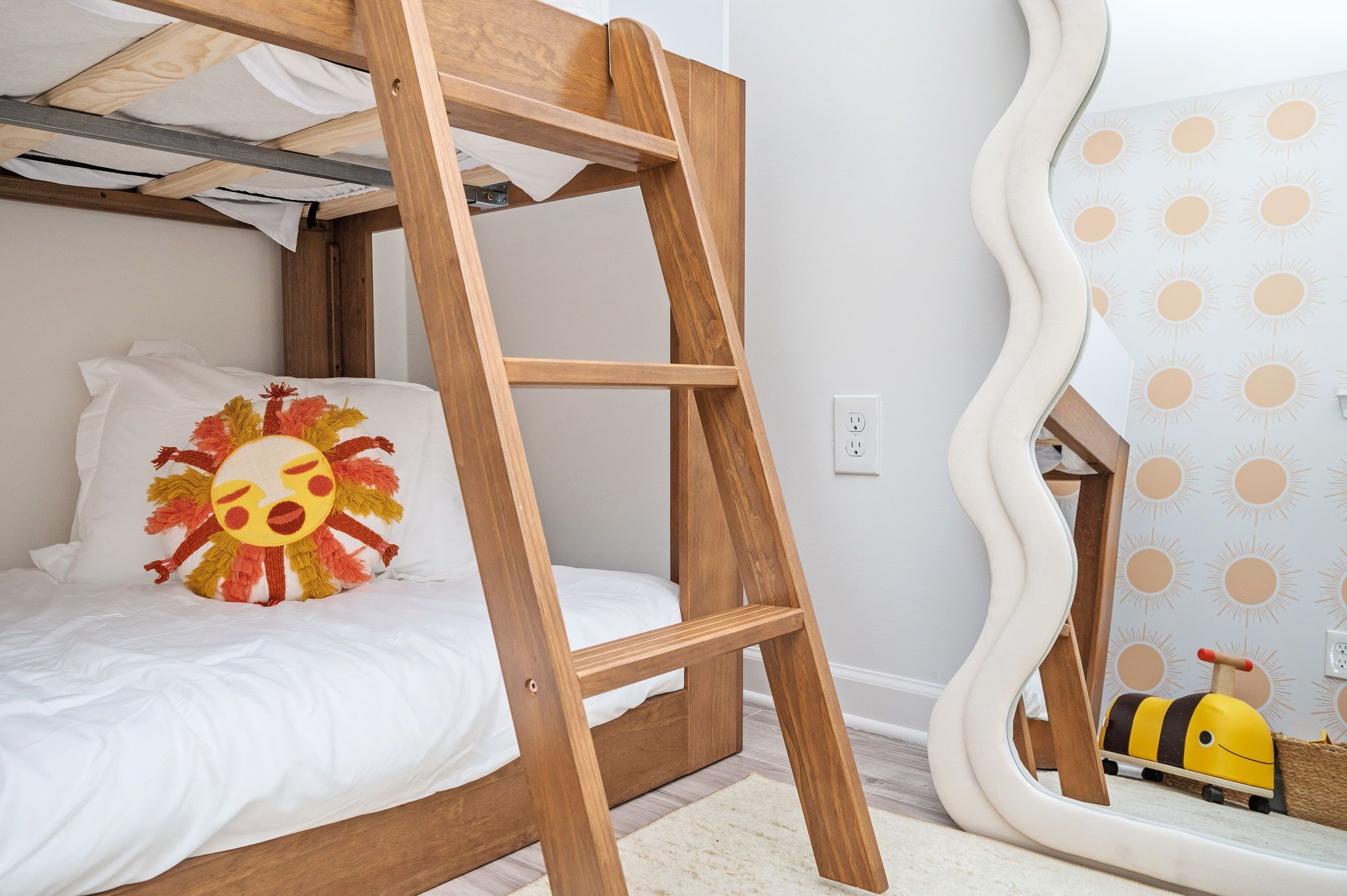 A bunk bed with a wooden ladder, a white pillow with a sun design, and a mirror reflecting a sun pattern wall.