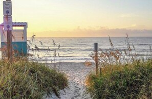 Une vue sur la plage avec un sentier menant à un bâtiment où est affiché un panneau indiquant un programme d'adoption de plages.