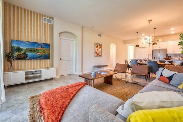 A modern living room with a sofa, coffee table, and TV. There is a kitchen area with cabinets and a dining table.