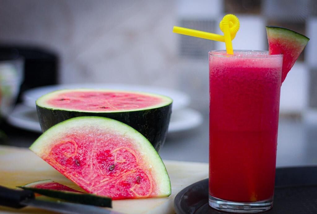 A glass of red beverage with a yellow straw and a slice of watermelon on the rim.