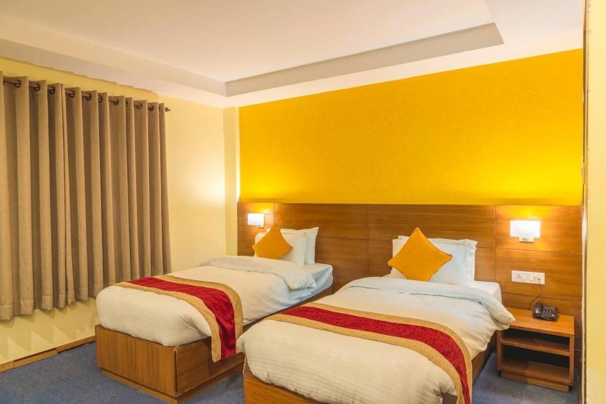 A hotel room with two beds, a wooden headboard, a nightstand, and a wall-mounted lamp.