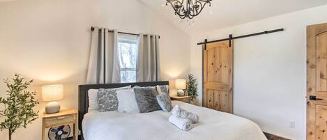 A bedroom with a large bed, a chandelier, a bedside table with a lamp, a fan, and a potted plant.