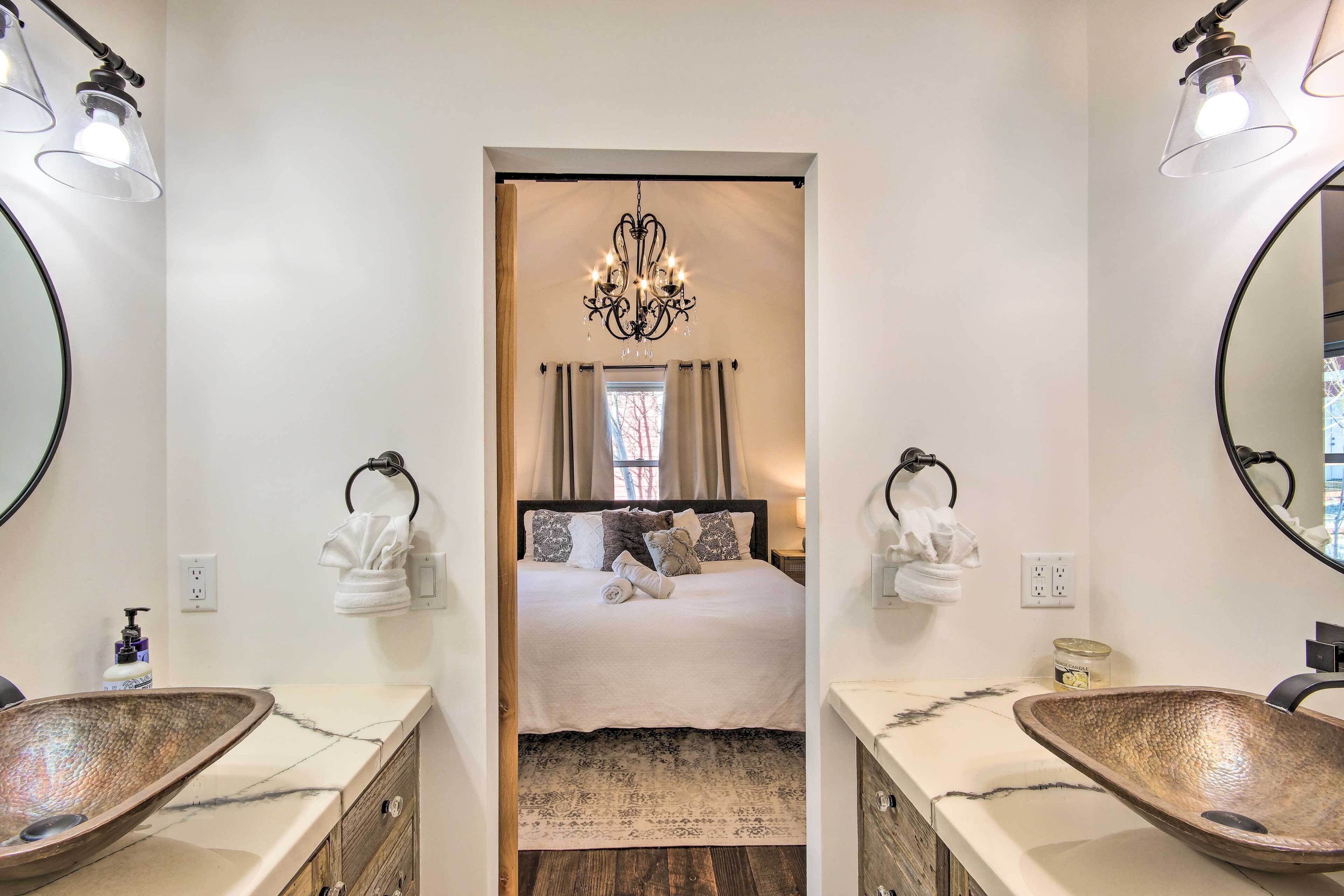 A hotel room with a large bed, two bedside tables with lamps, a chandelier, a large mirror, and a unique wooden sink.