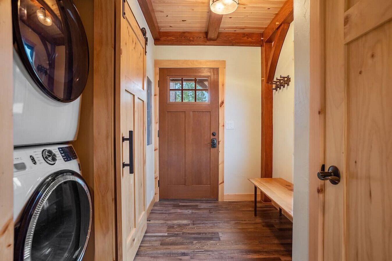 A laundry room with a washing machine and dryer, a wooden door with a window, and a bench in a hallway with wooden flooring.