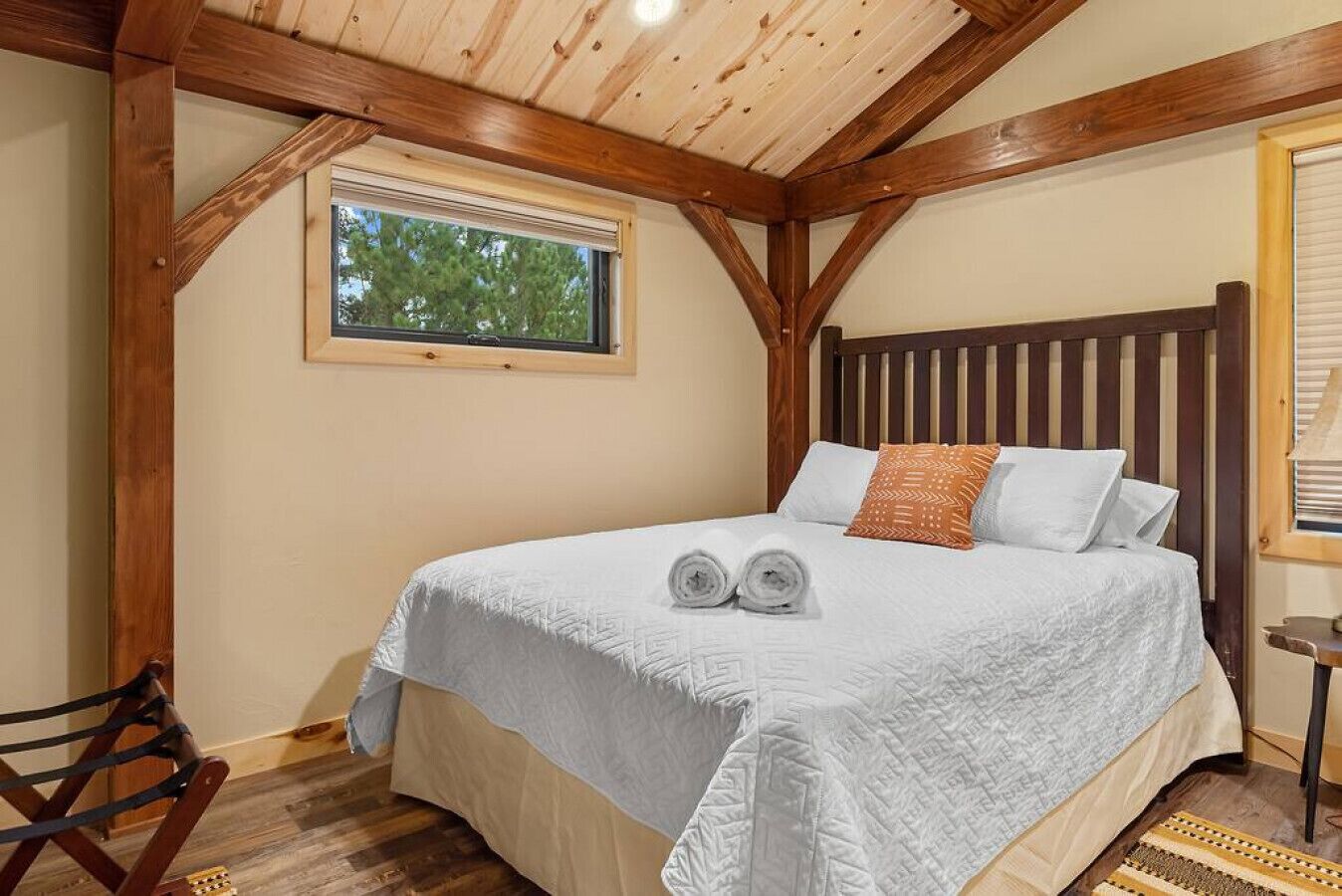 A bedroom with a wooden ceiling, a large bed with white bedding, a window, and a small table with a lamp.