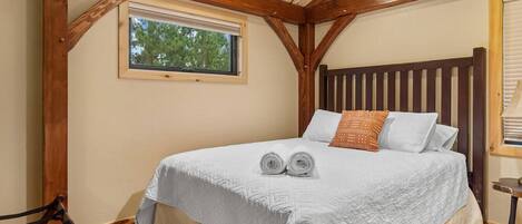 Un dormitorio con techo de madera, una cama grande con ropa de cama blanca, una ventana y una pequeña mesa con lámpara.