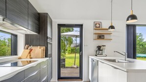 Een moderne keuken met donkere kastjes, een groot eiland en uitzicht op de tuin via een open deur.