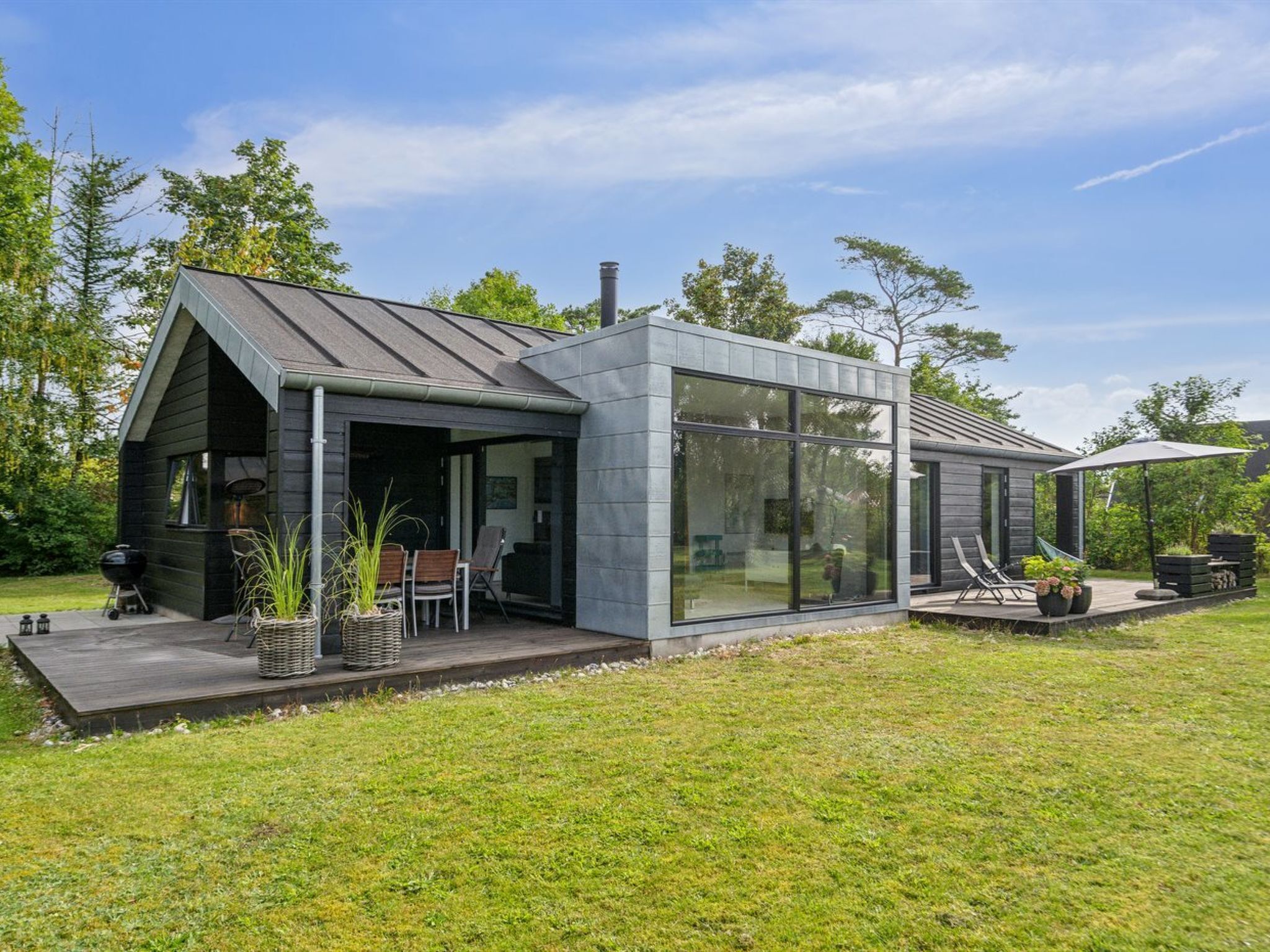 Ein modernes Haus mit einem großen Glasfenster, einer Terrasse mit Gartenmöbeln und einem Grillplatz.