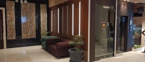 A modern lobby with a marble wall, a sofa, a potted plant, and a glass elevator.