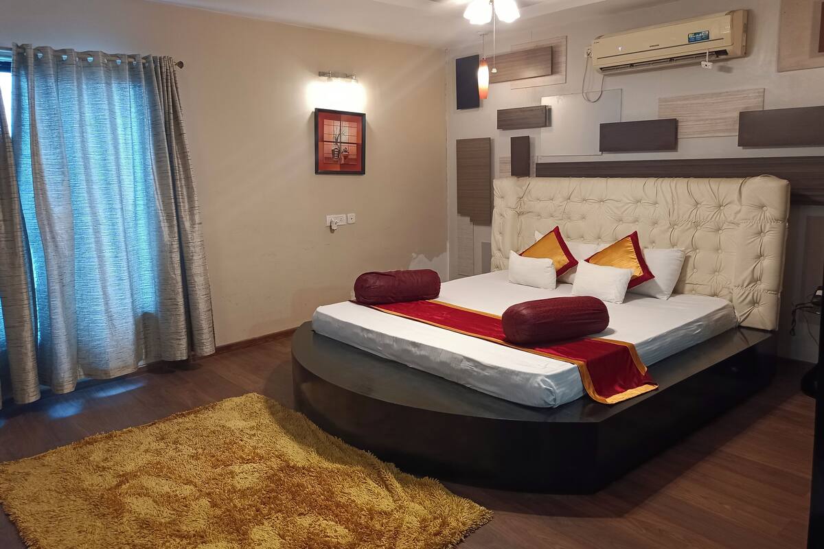 A modern bedroom with a tufted headboard, a large bed with red and white bedding, a yellow rug, and an air conditioning unit on the wall.