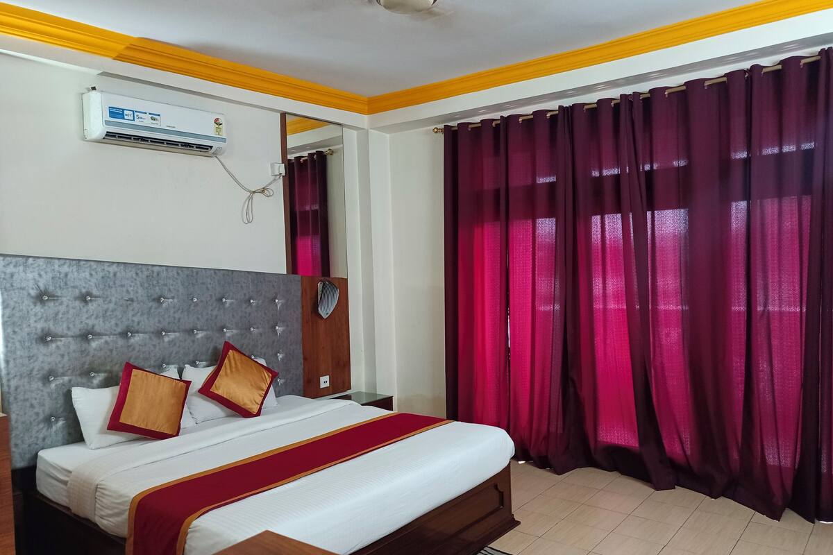 A hotel room with a bed, a bedside table, a mirror, a window with curtains, and an air conditioning unit.