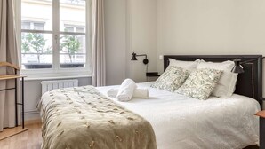 En pænt redt seng med hvid dyne og blomstrede puder, et natbord med en lampe, og et vindue med gardiner.
