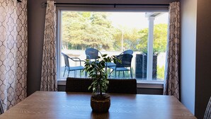 Uma área de jantar com uma mesa de madeira, um vaso com planta e vista para cadeiras e vegetação ao ar livre através de uma grande janela.
