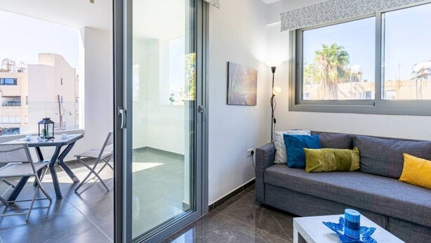 A modern living room with a grey sofa, a small table, and a glass door leading to a balcony. - Kmeidos 1-Bedroom Apartment & Balcony in Larnaca (Cyprus - Larnaca)