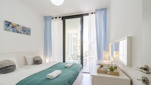 A modern hotel room with a bed, a bedside table with a tray of items, a window with curtains, and a view of the outside. - Lana 1-Bedroom Apt & Balcony in Larnaca (Cyprus - Larnaca)