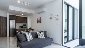 A modern living room with a grey sofa, a dining area, and a kitchen in the background. - Lana 1-Bedroom Apt & Balcony in Larnaca (Cyprus - Larnaca)