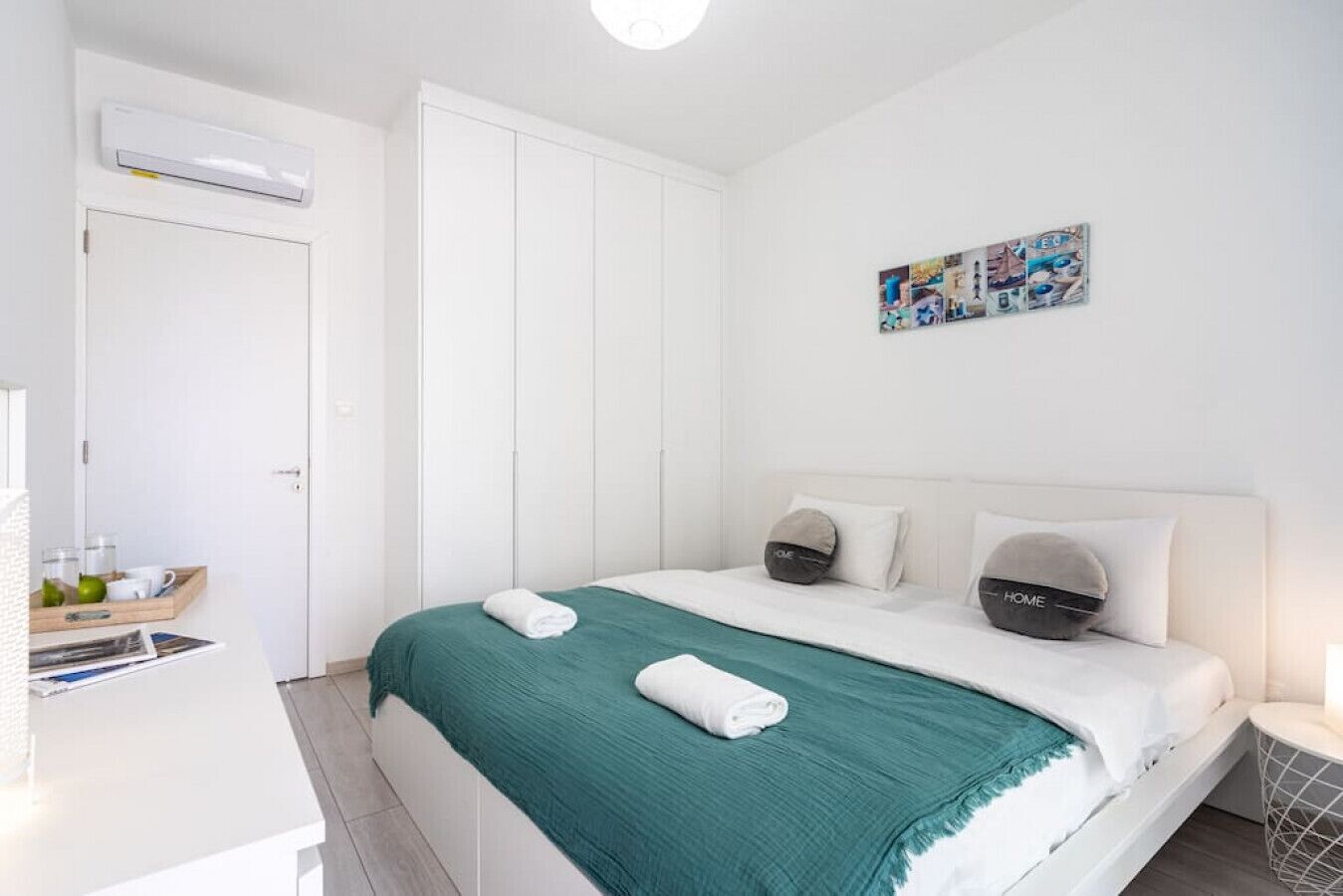 Quarto de hotel moderno com cama, mesa de cabeceira, ar condicionado fixado na parede e um pequeno estante com produtos de higiene pessoal.