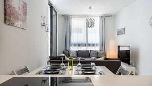 A modern living room with a sofa, a dining table set for four, and a large window with curtains. - Lana 1-Bedroom Apt & Balcony in Larnaca (Cyprus - Larnaca)