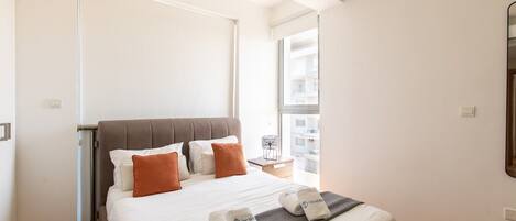 A modern hotel room with a large bed, a window with a city view, and a door leading to a balcony.