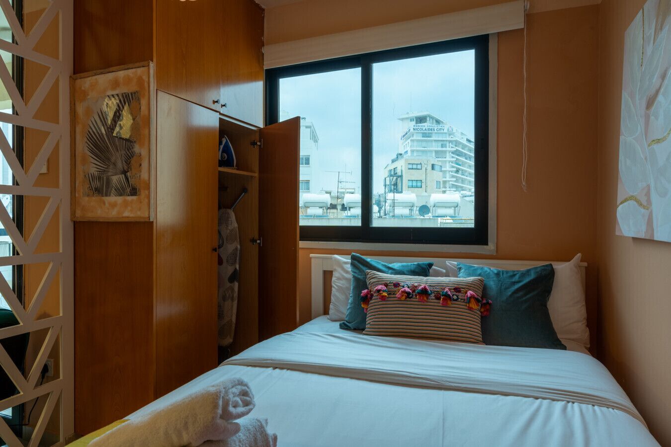 A bedroom with a bed, pillows, a window with a view of buildings, and a wooden wardrobe.