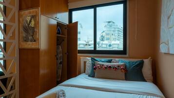 A bedroom with a bed, pillows, a window with a view of buildings, and a wooden wardrobe.