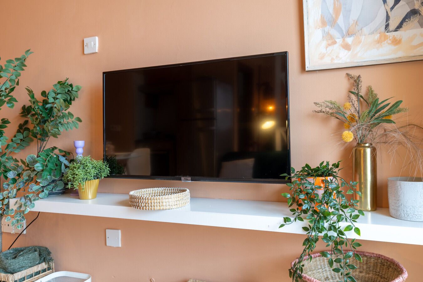 En moderne stue med et fladskærms-tv monteret på væggen, omgivet af planter og dekorative genstande på et hvidt konsolbord.