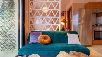 A teal sofa with patterned cushions, a wooden coffee table, and a kitchen area with a stainless steel refrigerator and oven.