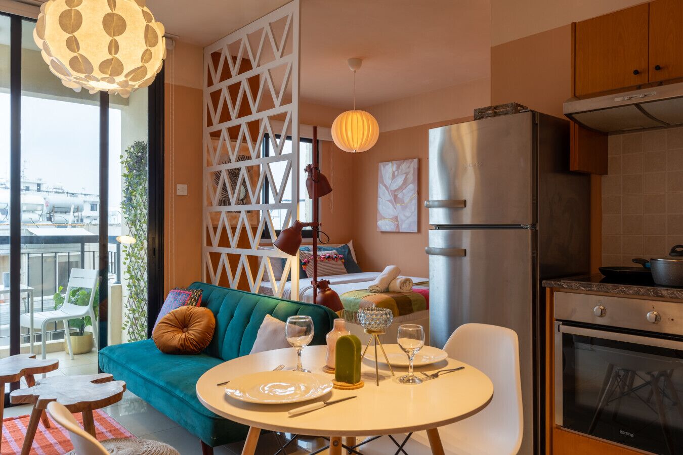 A modern living room with a teal sofa, dining table set with plates and glasses, a kitchen area with a stainless steel refrigerator, and a balcony with a view of the cityscape.