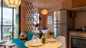 A modern living room with a teal sofa, dining table set with plates and glasses, a kitchen area with a stainless steel refrigerator, and a balcony with a view of the cityscape.