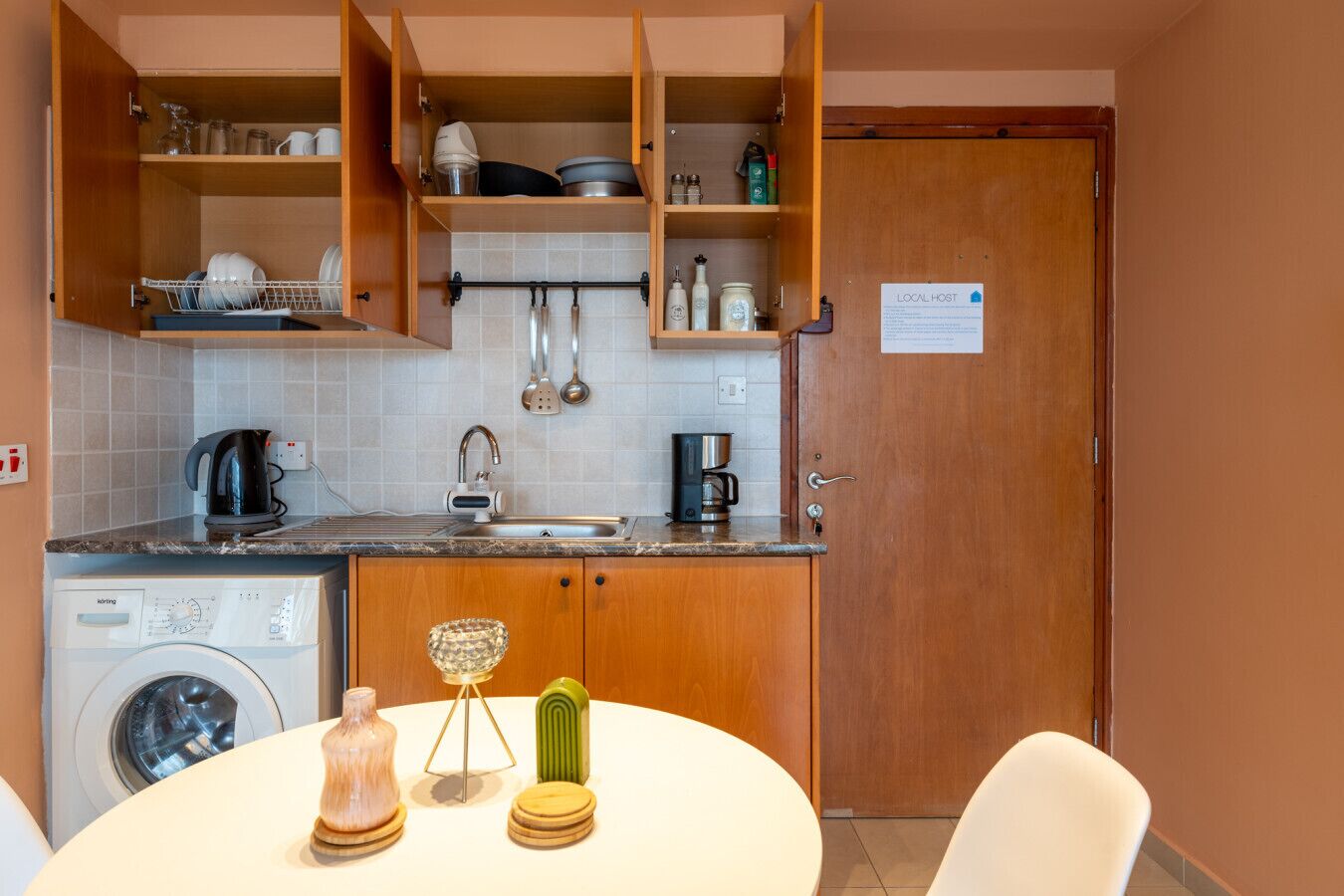Une cuisine compacte avec des meubles en bois, un lave-linge encastré, une machine à café et un panneau indiquant « HÔTE LOCAL ».