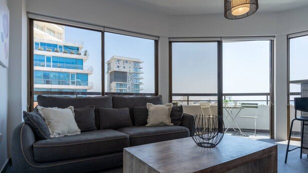 A modern living room with a dark grey sectional sofa, a wooden coffee table, and large windows offering views of buildings and a balcony. - Fiesta 1-Bedroom Apartment in Larnaca (Cyprus - Larnaca)