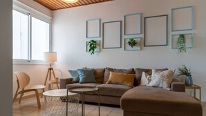 Ein Wohnzimmer mit einer Couch, einem Couchtisch, einem Sessel und einer Lampe. An den Wänden hängen Bilderrahmen und auf dem Nachttisch steht eine Pflanze.