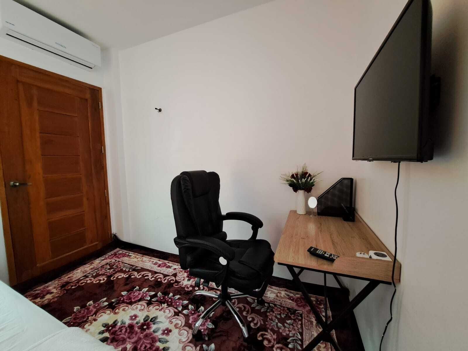 Una habitación pequeña con una puerta de madera, una silla de oficina negra, una pequeña mesa de madera con un control remoto, un florero con flores y un televisor montado.