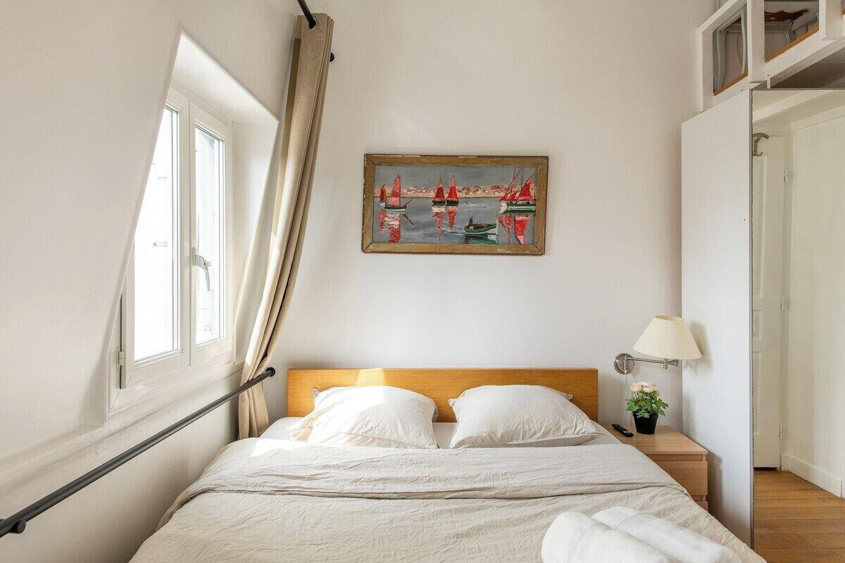 A bedroom with a bed, a nightstand, a lamp, a painting, and a window with curtains.