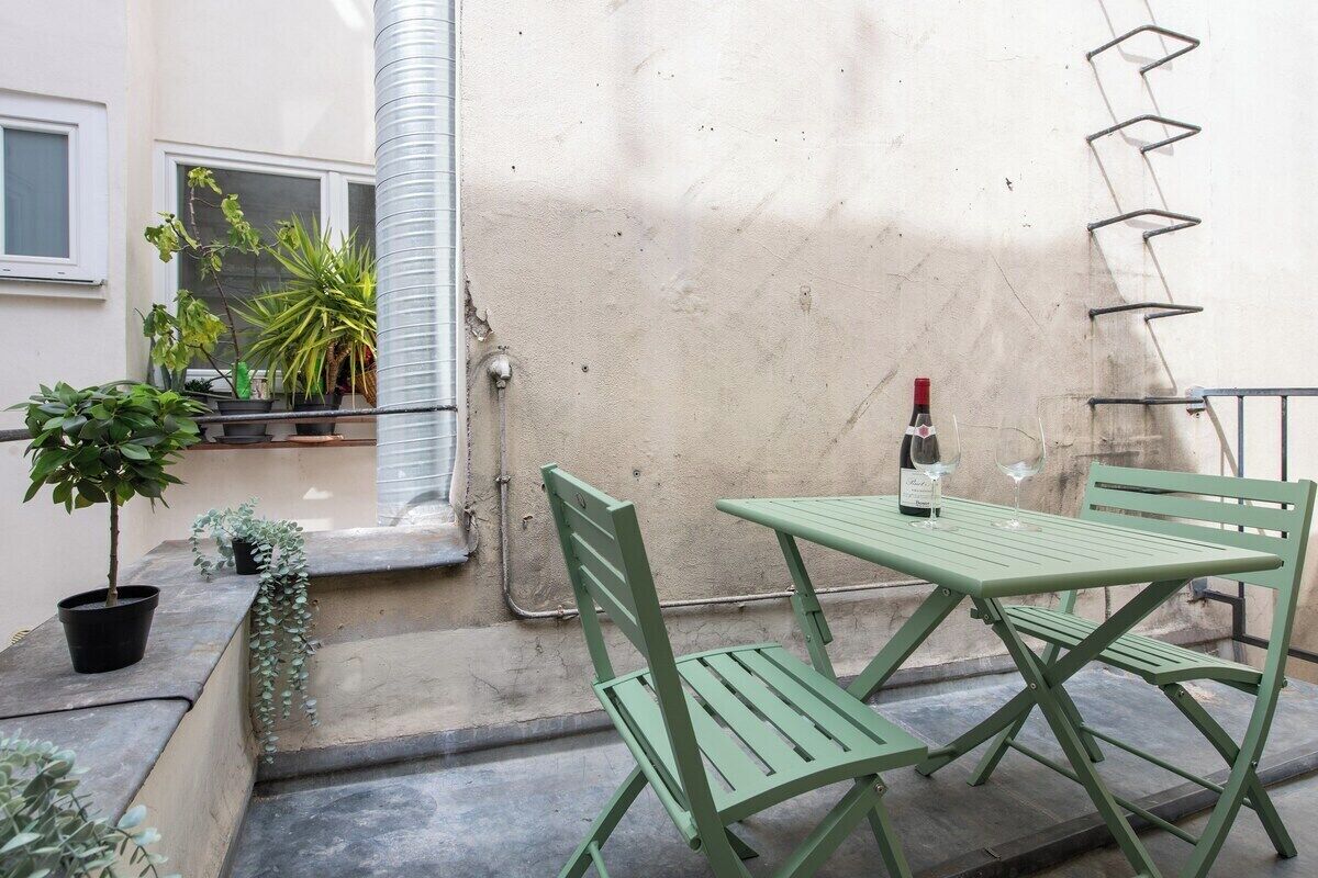 A small outdoor patio with a green metal table and chairs, a bottle of wine, and potted plants.