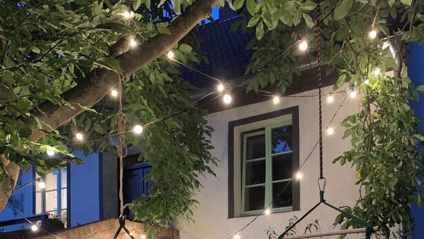 A hammock under a tree with string lights, a chair, and a brick wall.