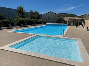 Una piscina rectangular con un trampolín, rodeada de sillas de descanso y un edificio con techo de tejas.