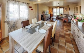 A dining area with a wooden table set for four, a kitchen in the background, and a living area with a sofa and a lamp.
