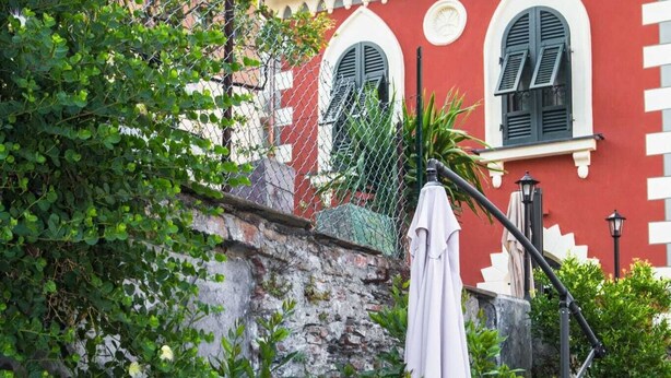 Un patio con mobiliario exterior, una sombrilla blanca y un edificio rojo con ventanas arqueadas al fondo.