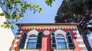 Un edificio con dos ventanas arqueadas con persianas verdes, una puerta blanca y un árbol al fondo.