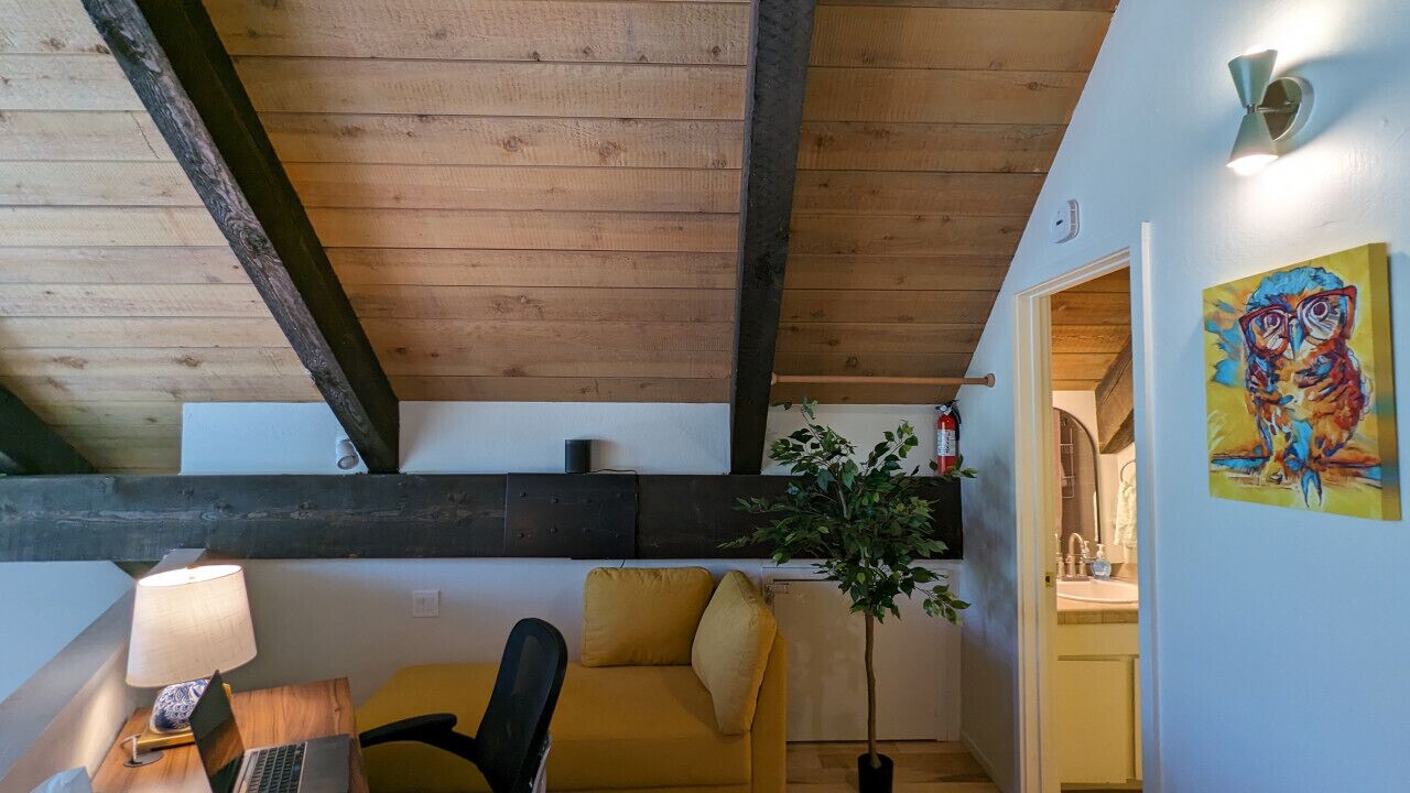 A cozy room with a wooden ceiling, a yellow sofa, a desk with a lamp, and a painting on the wall.