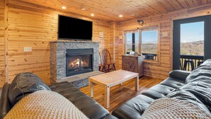 A cozy cabin interior with a stone fireplace, leather sofa, wooden walls, and a large window offering a scenic view. - Steps from Hatley Pointe - Short drive to downtown Asheville! (Mars Hill)
