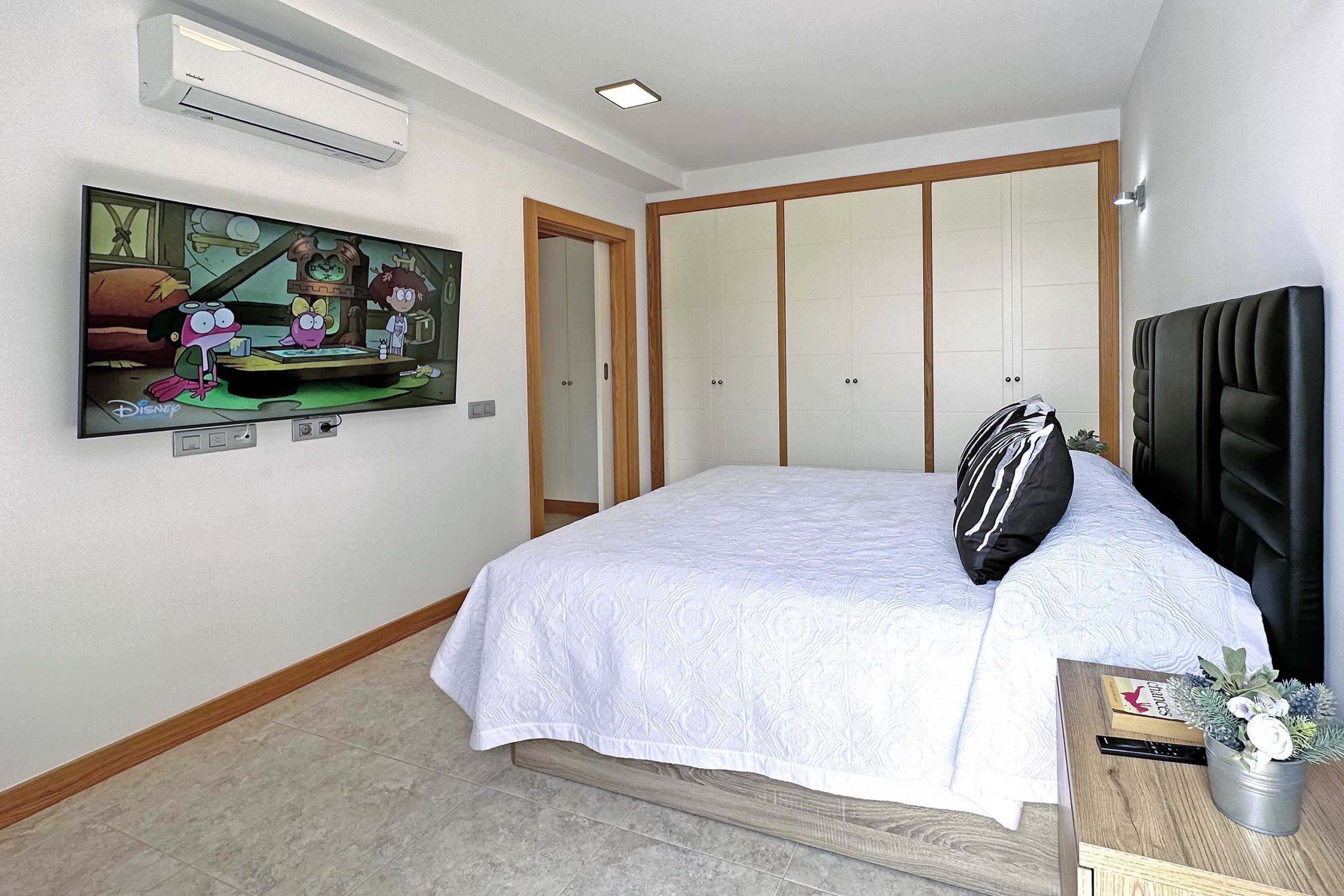 Un dormitorio con cama, televisor montado en la pared, armario y una mesita con una planta.