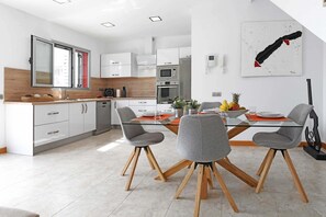 A modern kitchen with white cabinets, a built-in oven, and a dining area with a glass table and chairs. - Salobre Vista Golf 8 (Maspalomas)