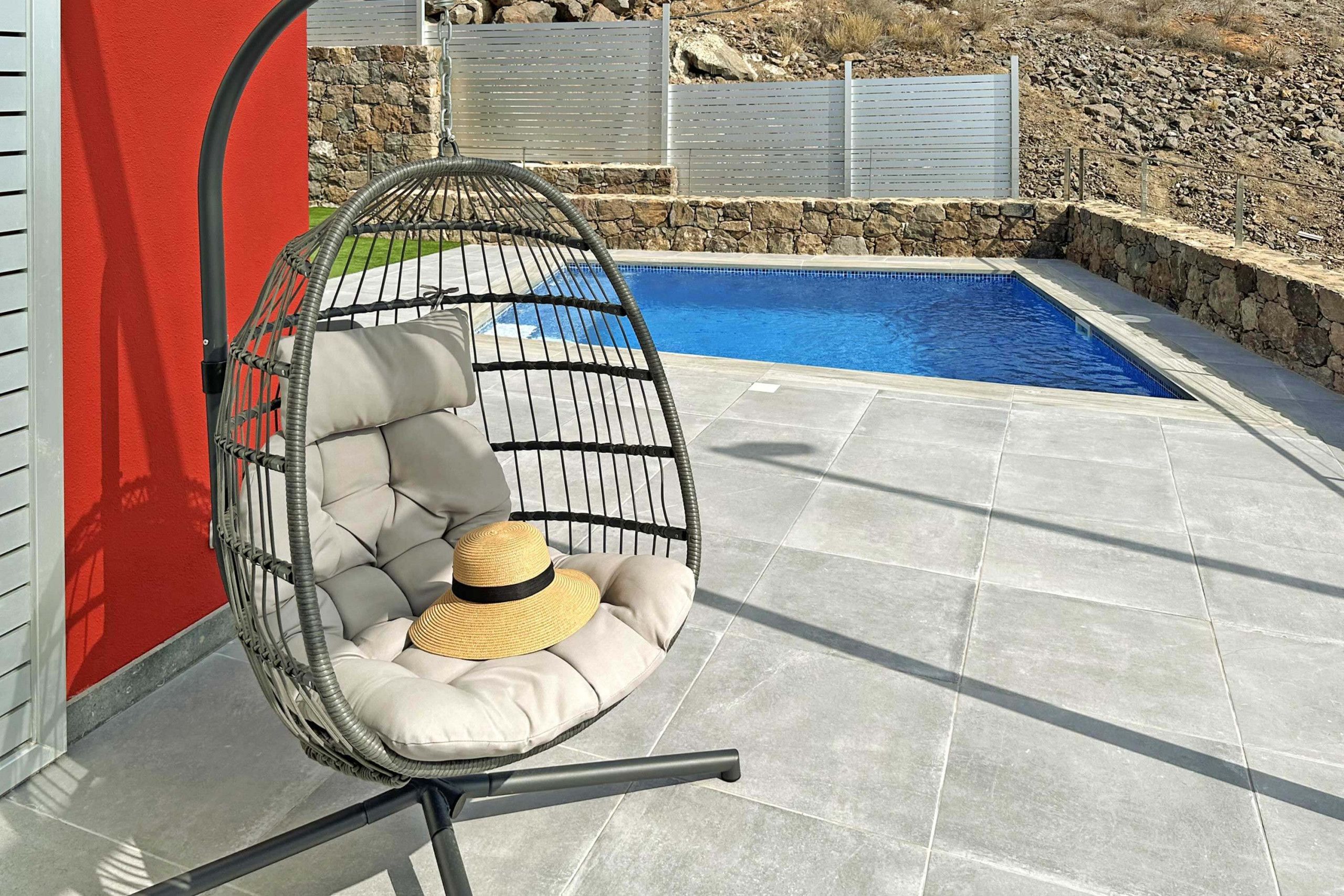 A hanging chair with cushions and a straw hat, positioned near a swimming pool with a stone wall and a metal fence in the background.