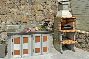 Cozinha externa com parede de pedra, pia e forno a pizza.